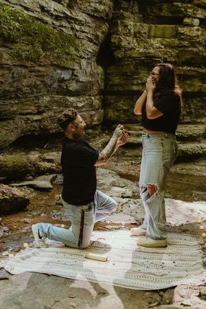 A couple sits on a blanket having a picnic near a rocky cliff wall in a natural outdoor setting.