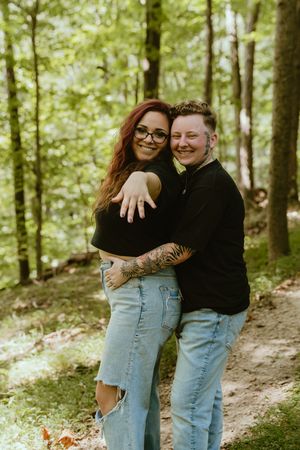 A couple in jeans and black tops share a tender embrace while standing on a wooded forest trail.