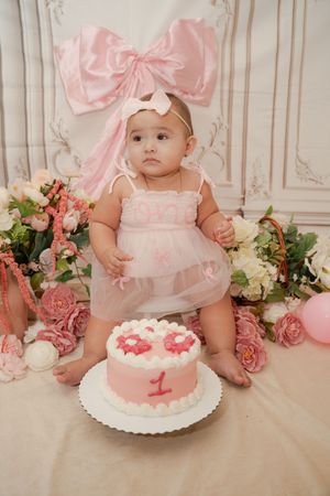 Baby girl in pink dress with bow headband sits beside a pink number 1 birthday cake surrounded by flowers.