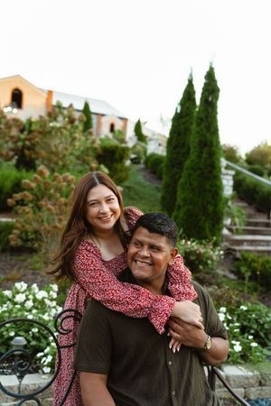 A couple embraces outdoors in a garden setting with cypress trees and flowers in the background.