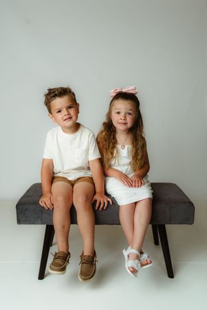 Two children sit together on a dark wooden bench wearing white outfits and contrasting shoes.