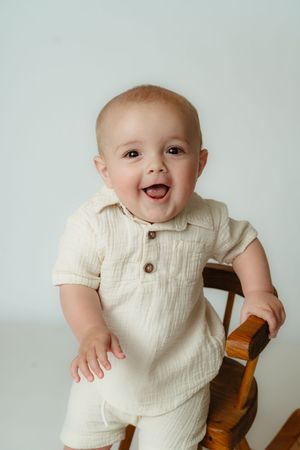 A happy baby in a cream-colored outfit sits in a wooden chair against a light background.