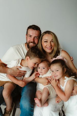 A family wearing coordinated white outfits poses together for a casual portrait session against a gray backdrop.