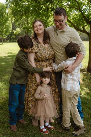Family wearing coordinated earth-toned outfits embraces in a sunny outdoor garden setting.