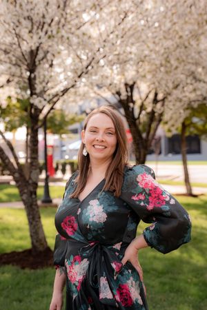 Woman in floral black dress smiling outdoors near blooming white cherry blossom trees on green lawn.
