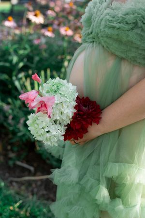 Pregnant woman in sage green tulle gown holding bouquet of white hydrangeas and red dahlias in garden.