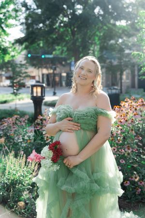 Pregnant woman in green off-shoulder tulle gown holding flowers, posing in a garden with colorful blooms.