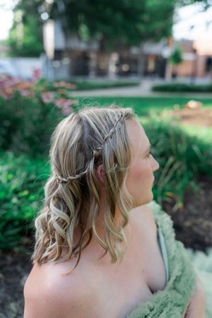 Woman with blonde braided half-up hairstyle and soft curls wearing a green dress in a garden setting.