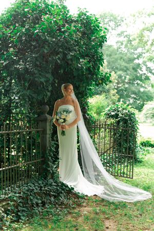 Bride in white strapless gown with long veil holds bouquet by ivy-covered iron garden gate.