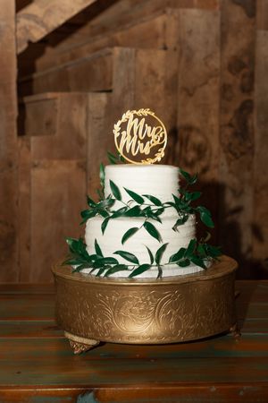 Two-tier white wedding cake with green leaf garland and gold Mr. & Mrs. topper on ornate gold stand.