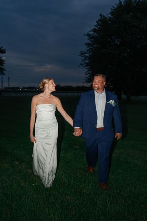 Bride and groom holding hands walking on grass at dusk, she in white gown, he in navy suit.
