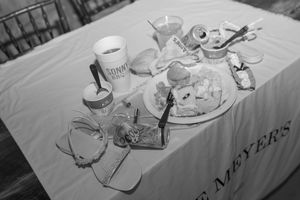 Black and white photo of a cluttered table with food, drinks, and utensils on a white tablecloth at an event.