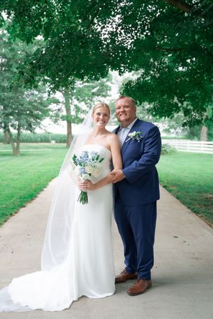Bride and groom pose on a tree-lined path, she holds a blue bouquet in a white gown, he wears a navy suit.