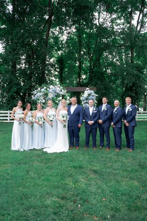 Wedding party poses outdoors in light blue bridesmaid dresses and navy suits near a floral arch.