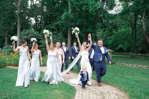 Wedding party celebrating outdoors on a garden path, bride and bridesmaids raising bouquets joyfully with groomsmen and a young child.
