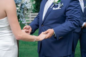 Bride and groom exchanging rings during outdoor wedding ceremony, groom wearing navy blue suit with floral boutonniere.