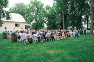 Outdoor wedding ceremony on green lawn with white chairs, barrel decor, and a white barn surrounded by tall trees.