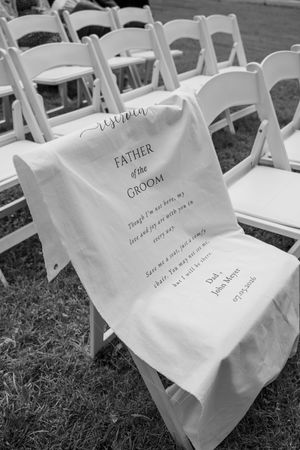 Black and white photo of a memorial chair sign honoring the Father of the Groom at an outdoor wedding ceremony.