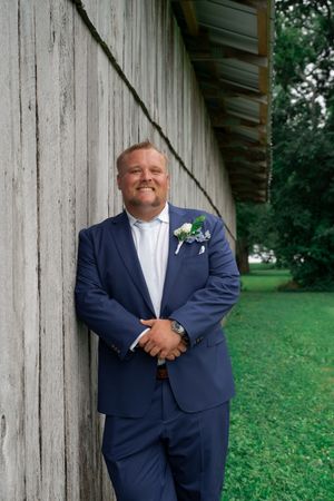 A smiling groom in a navy blue suit with boutonniere leans against a rustic wooden barn wall outdoors.