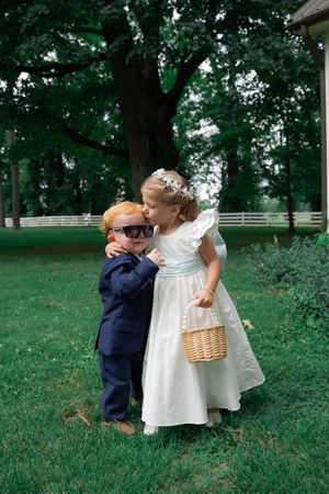 Adorable flower girl kisses ring bearer wearing sunglasses in navy suit on green lawn at summer wedding.