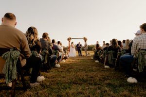 A rustic outdoor wedding ceremony at sunset with guests seated on the lawn facing a wooden arch.
