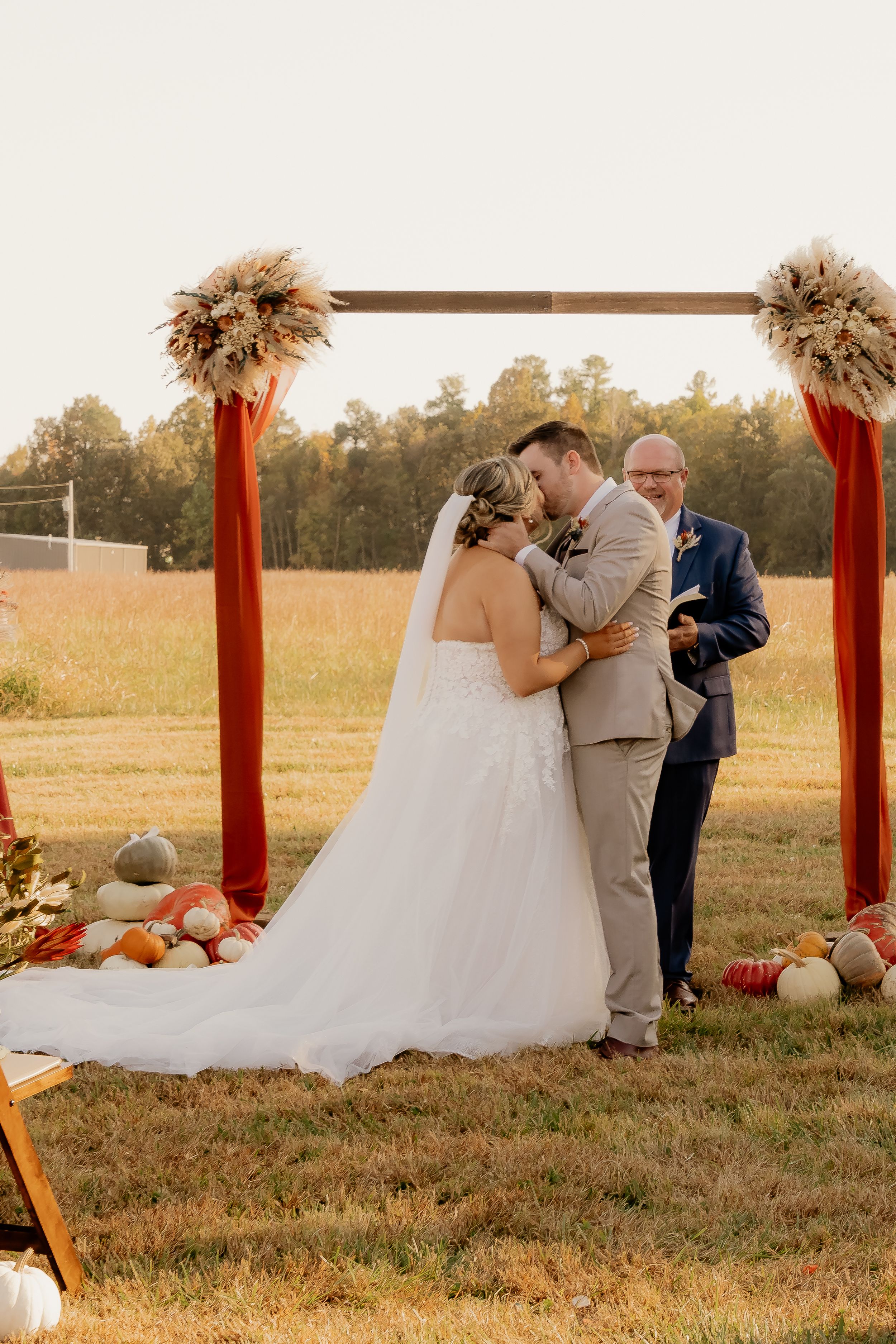 Fall wedding ceremony under wooden arch decorated with dried pampas grass and pumpkins.