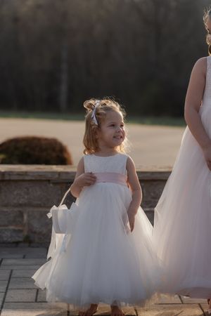 A smiling flower girl in a white tulle dress with pink sash carries a basket at an outdoor wedding ceremony.