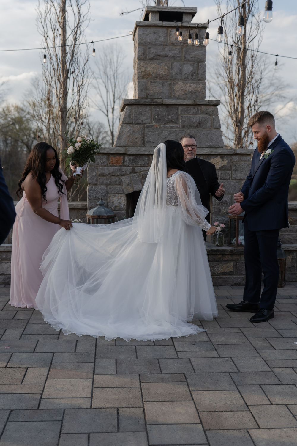 Bride in white gown and veil at outdoor wedding ceremony by stone fireplace with groom and bridesmaid.