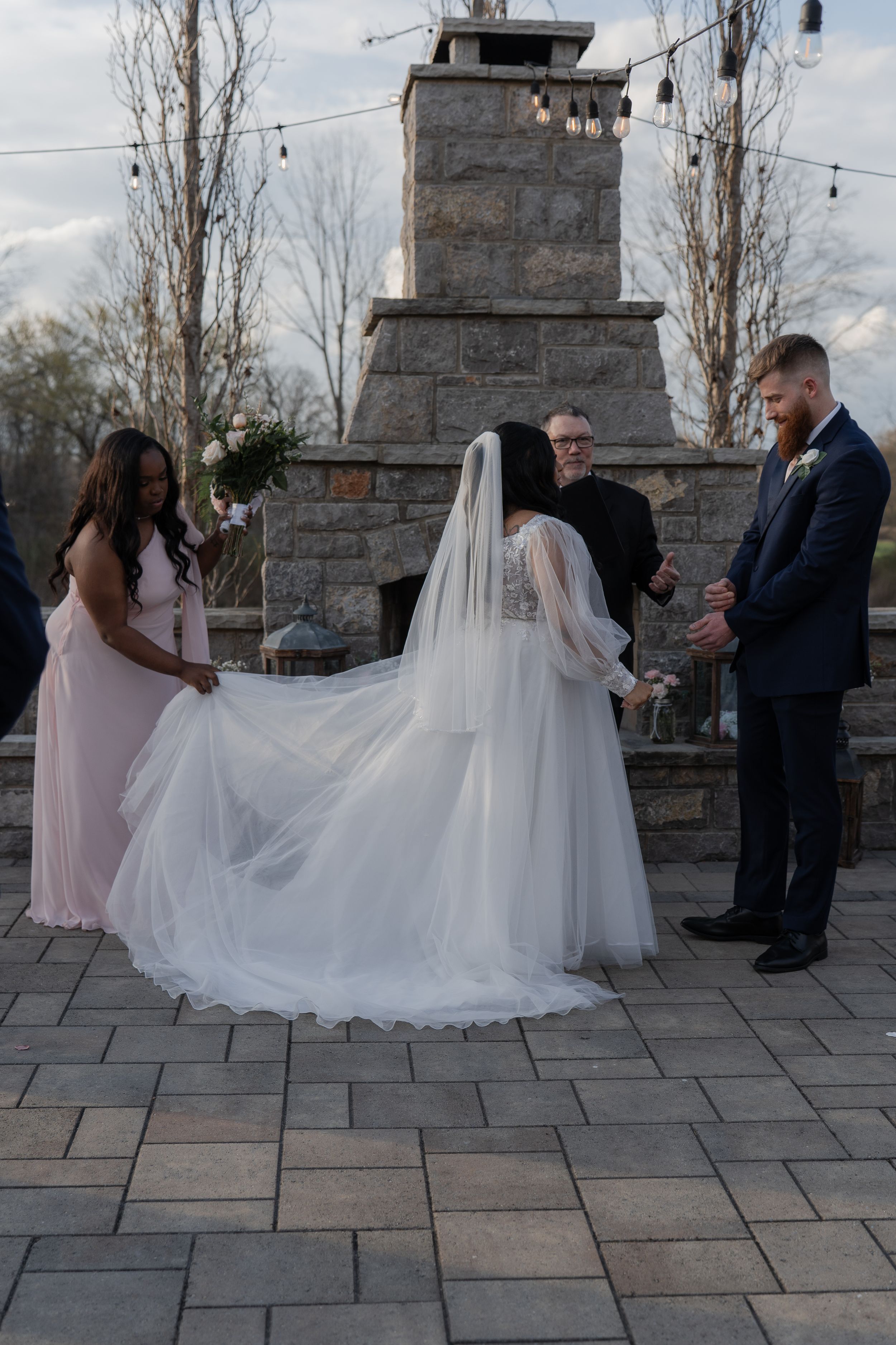 Bride in white gown and veil at outdoor wedding ceremony by stone fireplace with groom and bridesmaid.