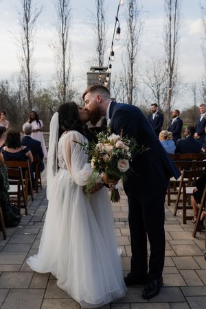 Bride and groom share first kiss at outdoor wedding ceremony on stone patio with string lights and guests seated behind them.