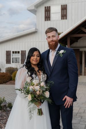 Bride in white lace gown holding floral bouquet poses with groom in navy suit outside white barn venue.