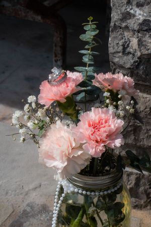 Pink carnations and baby's breath in a mason jar with pearl beads and wedding rings, set against a stone wall backdrop.