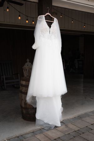 White lace wedding dress with veil hanging on a wooden hanger in a rustic barn venue with string lights.