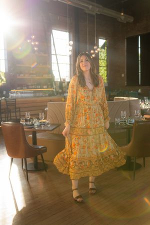 A person in a flowing yellow floral dress poses in a sunlit restaurant interior with wooden floors and elegant seating.