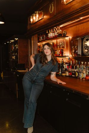 A retro-style bar interior with warm lighting, wooden decor, and a denim-clad figure leaning against the counter.