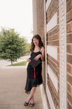 Pregnant woman in black dress holding graduation cap with red tassel, leaning against brick wall outdoors.