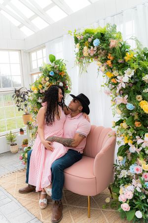Couple in matching pink outfits sitting on a pink chair under a floral arch in a bright greenhouse setting.