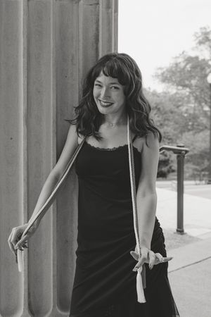 Black and white photo of a smiling woman in a black slip dress holding a rope, leaning against a large stone column outdoors.