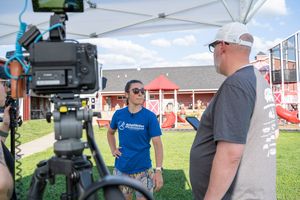 Two men talking under a tent at an outdoor event, with a camera on a tripod in the foreground and a red barn behind.