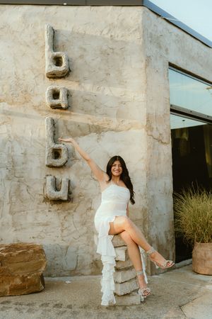 A person in a white dress poses next to a vertical 'buu' sign mounted on a textured concrete wall.