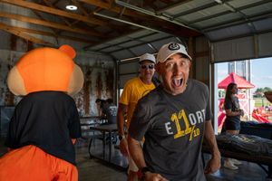 Man in gray shirt laughing near orange mascot costume at outdoor event with friends in background.