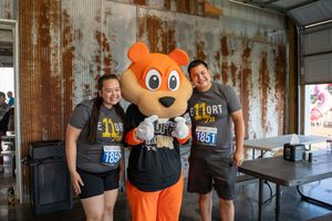 Two runners pose with an orange bear mascot at an outdoor charity run event, wearing matching gray race t-shirts.