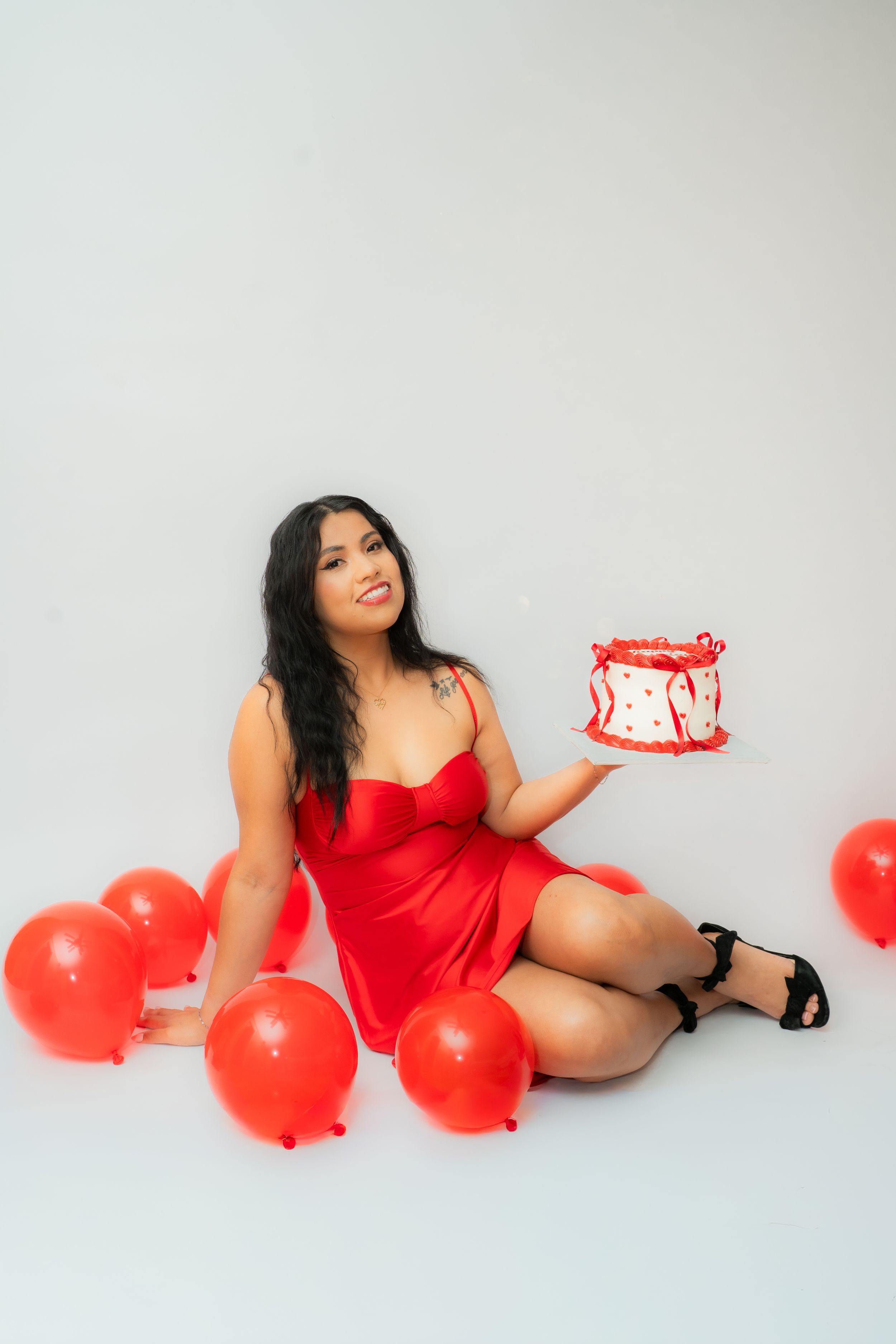 Woman in red dress sitting on floor holding decorated cake surrounded by red balloons on white background.