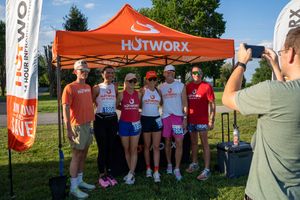Group of runners posing under an orange HOTWORX tent at an outdoor race event on a sunny day.