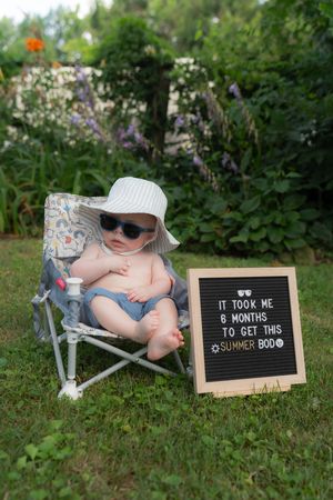 Cute 6-month-old baby in sunglasses and sun hat sitting in mini chair outdoors next to a funny summer bod letter board.