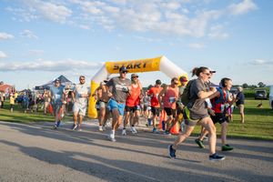 Runners cross the start line at an outdoor 5K race on a sunny morning with a yellow inflatable arch.