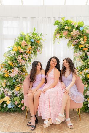 Three women in pastel dresses pose on a pink velvet sofa surrounded by a lush floral arch with colorful blooms.