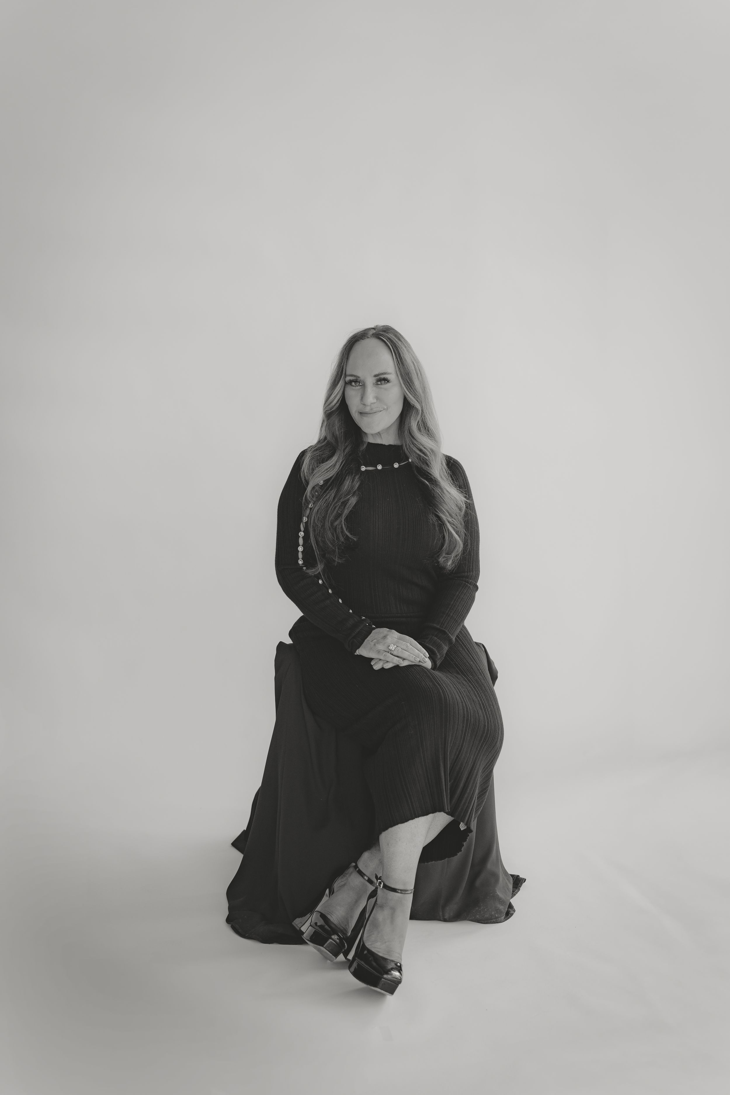 Woman in black maxi dress and heels seated elegantly in a minimalist black and white studio portrait.