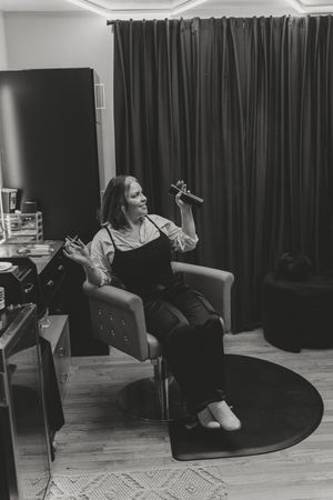 Black and white photo of a woman sitting in a chair holding makeup brushes in a stylish dressing room.