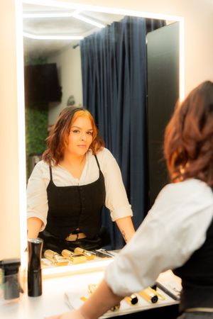 Woman in black apron examines her reflection in a lit vanity mirror at a professional makeup station.